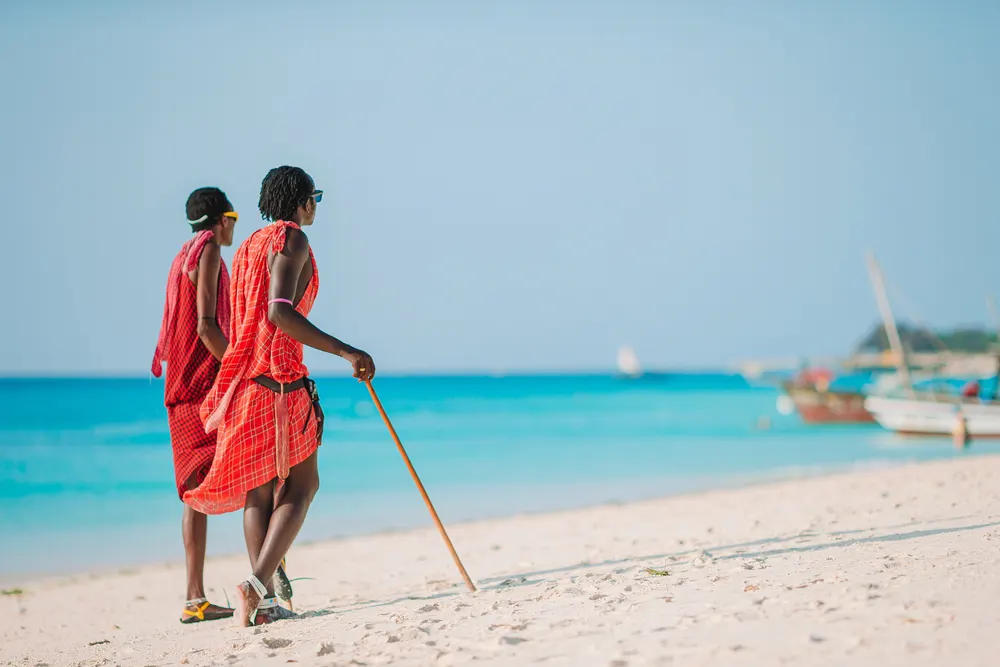 Two Maasai men in red shuka robes walking along a white-sand Zanzibar beach with wooden dhow boats offshore.
